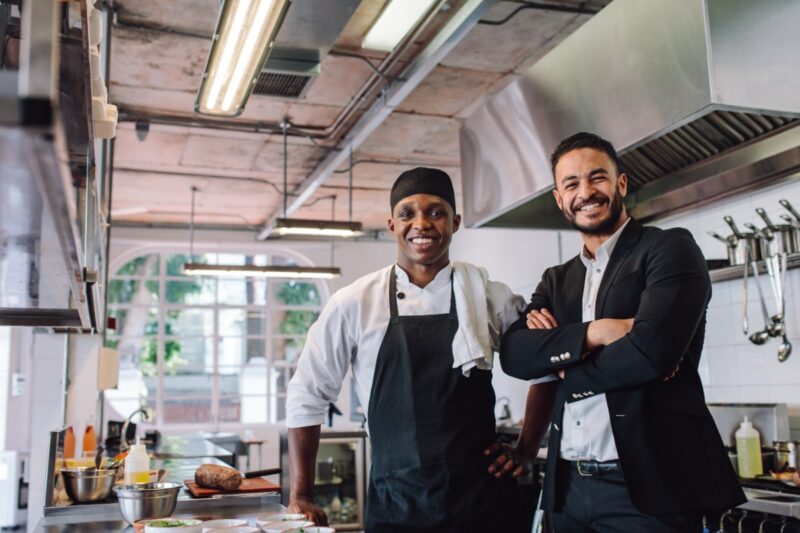 Two black men, one in suit and one in chef shirt and apron, smiling inside commercial kitchen